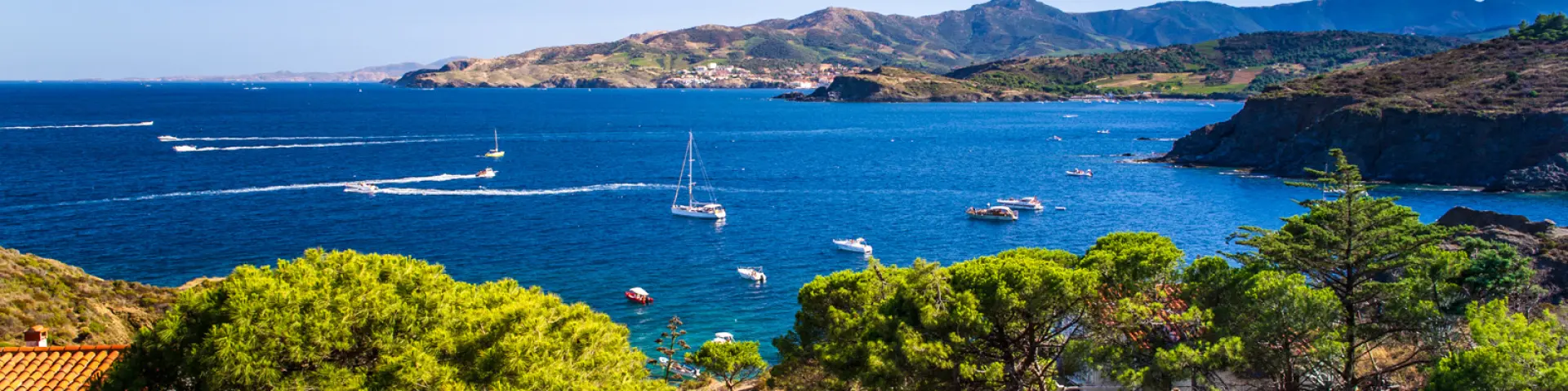 Vue panoramique sur la Côte Vermeille depuis les hauteurs, avec la mer Méditerranée d’un bleu profond, des voiliers au mouillage et les collines verdoyantes plongeant dans la mer.