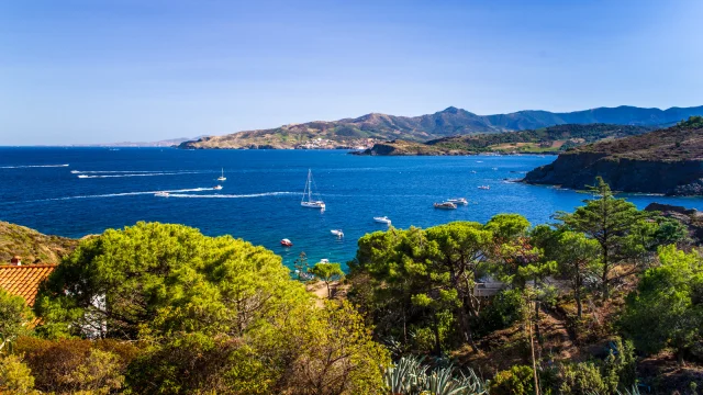 Vue panoramique sur la Côte Vermeille depuis les hauteurs, avec la mer Méditerranée d’un bleu profond, des voiliers au mouillage et les collines verdoyantes plongeant dans la mer.