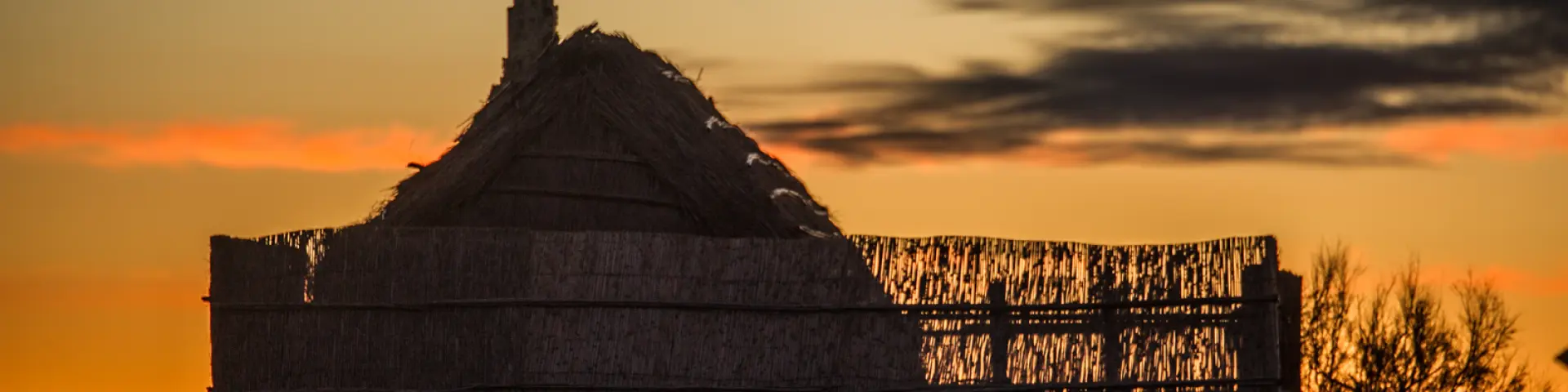 Cabane traditionnelle en roseaux silhouettée au coucher du soleil, sur fond de ciel doré et orangé.