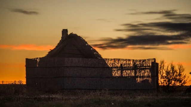 Cabane traditionnelle en roseaux silhouettée au coucher du soleil, sur fond de ciel doré et orangé.
