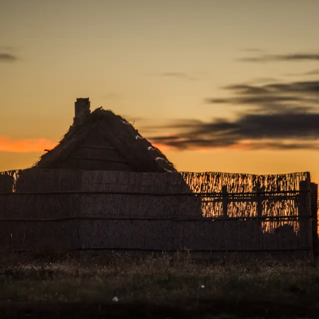 Cabane traditionnelle en roseaux silhouettée au coucher du soleil, sur fond de ciel doré et orangé.