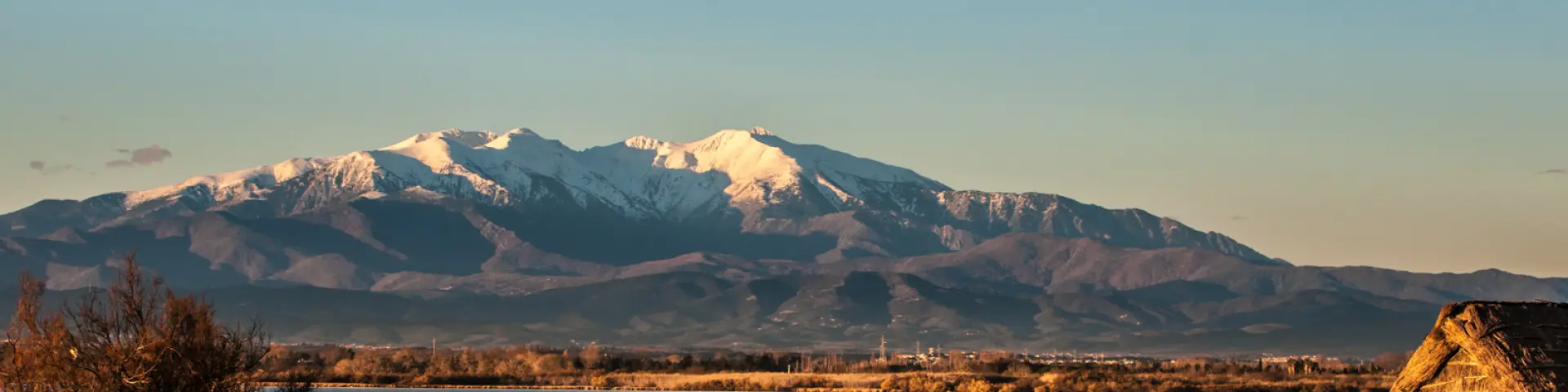 Vue sur une cabane traditionnelle en roseaux au bord d’un étang, avec en arrière-plan le Canigó enneigé sous la lumière dorée du soir.