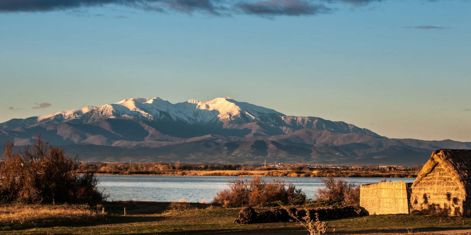 Vue sur une cabane traditionnelle en roseaux au bord d’un étang, avec en arrière-plan le Canigó enneigé sous la lumière dorée du soir.