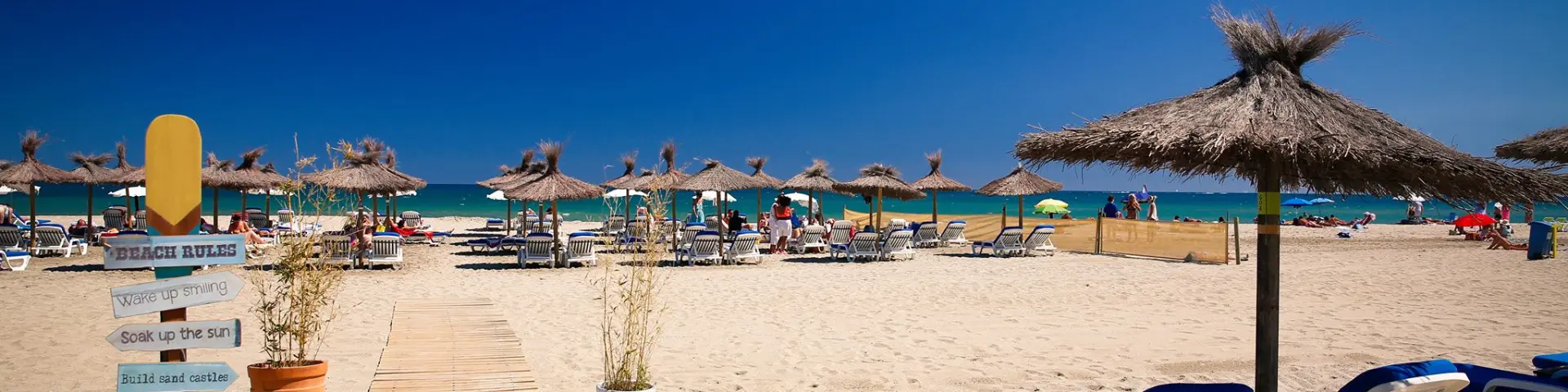 Plage aménagée de sable fin à Canet-en-Roussillon, avec transats, parasols en paille et promenade en bois menant jusqu’à la mer sous un ciel bleu éclatant.