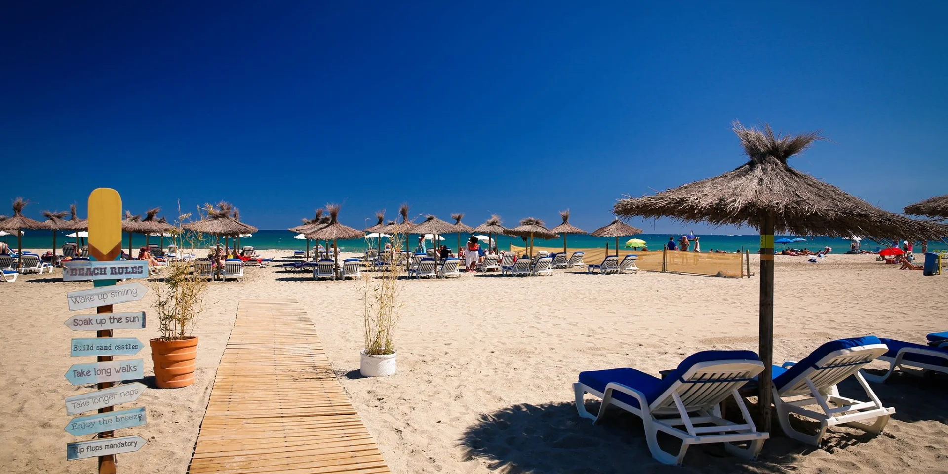 Plage aménagée de sable fin à Canet-en-Roussillon, avec transats, parasols en paille et promenade en bois menant jusqu’à la mer sous un ciel bleu éclatant.