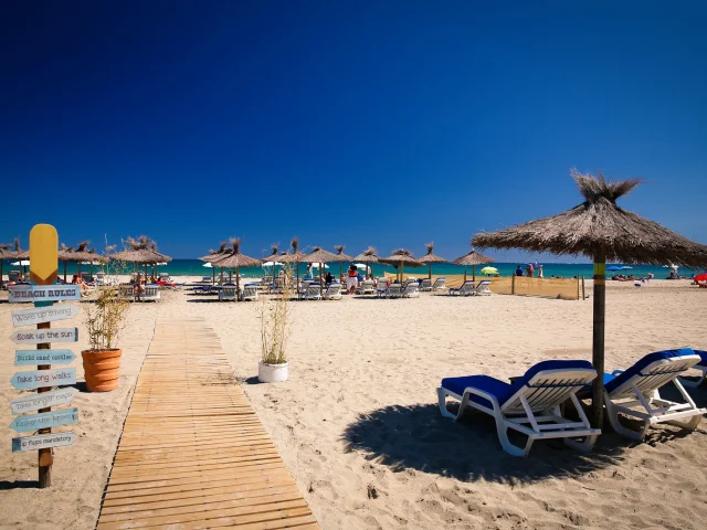 Plage aménagée de sable fin à Canet-en-Roussillon, avec transats, parasols en paille et promenade en bois menant jusqu’à la mer sous un ciel bleu éclatant.