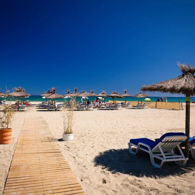 Plage aménagée de sable fin à Canet-en-Roussillon, avec transats, parasols en paille et promenade en bois menant jusqu’à la mer sous un ciel bleu éclatant.