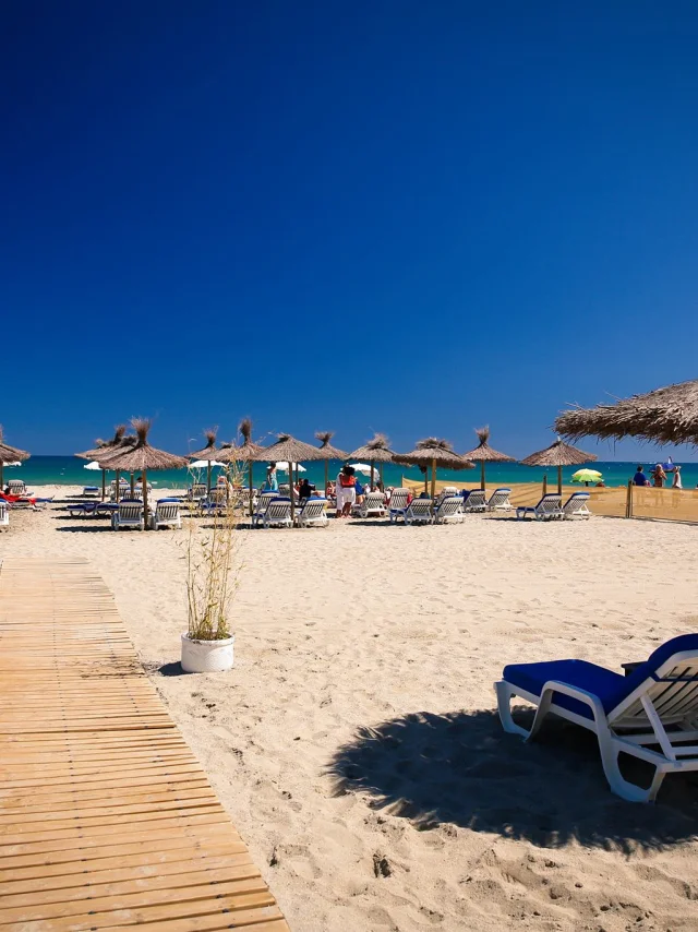 Plage aménagée de sable fin à Canet-en-Roussillon, avec transats, parasols en paille et promenade en bois menant jusqu’à la mer sous un ciel bleu éclatant.