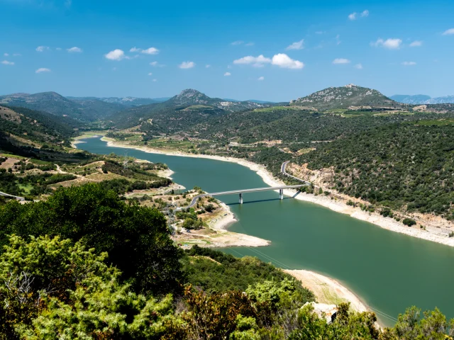 Vue panoramique sur le lac de l’Agly entouré de collines verdoyantes, avec un pont traversant les eaux calmes sous un ciel bleu.
