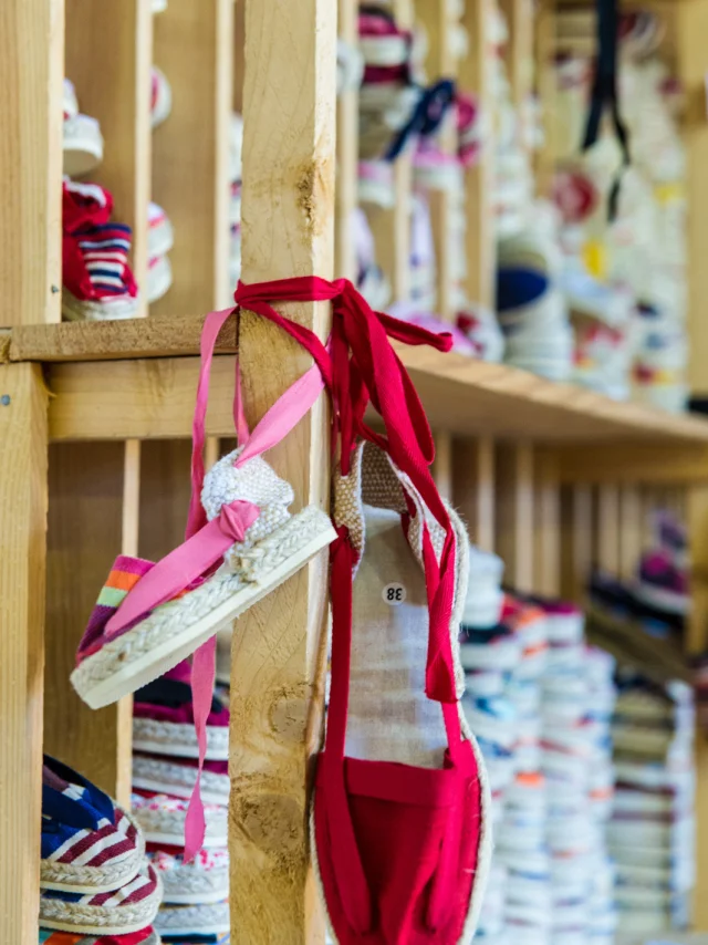Paire d’espadrilles rouges accrochée à une étagère en bois dans un atelier de fabrication, entourée d’autres modèles colorés empilés.