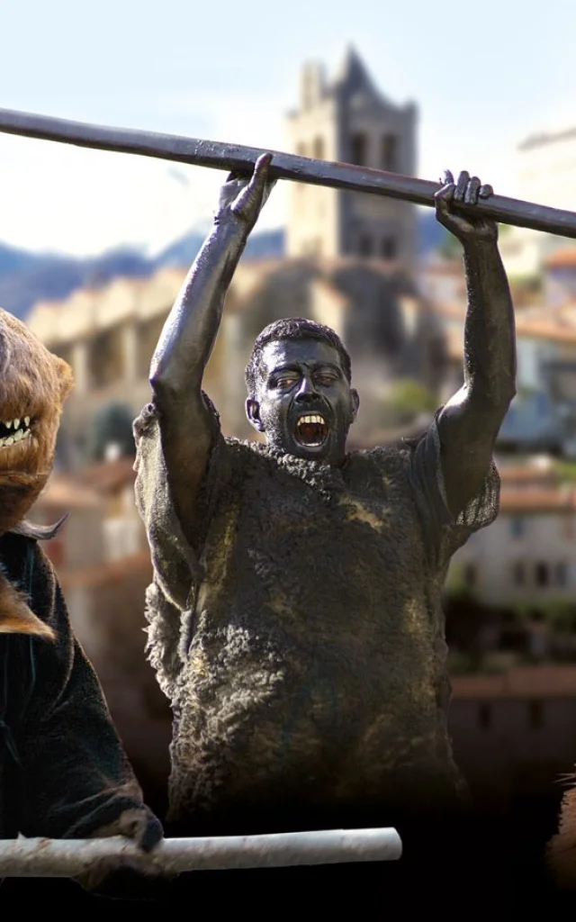Personnages costumés incarnant la Fête de l’Ours dans un village des Pyrénées, avec des participants grimés et des costumes d’ours sur fond de clochers et de montagnes.