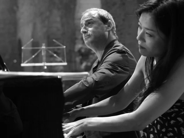Deux pianistes jouant à quatre mains sur scène lors d’un concert, en noir et blanc.