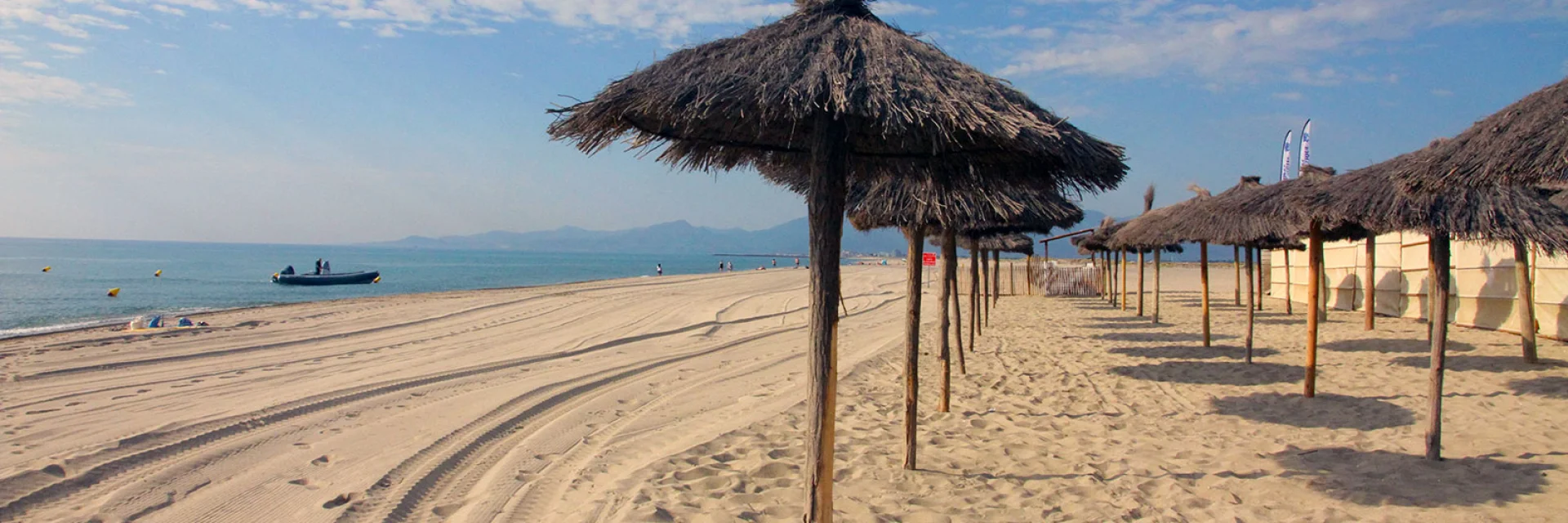 Plage de sable fin bordée de parasols en paille, avec la mer Méditerranée et les montagnes visibles à l’horizon sous un ciel bleu.