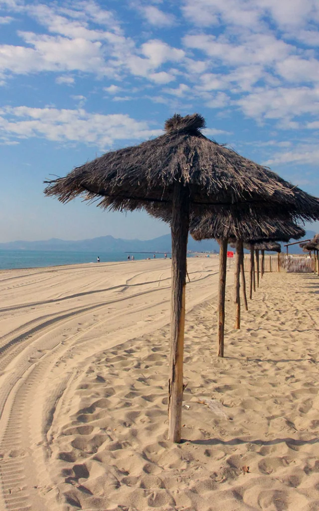 Plage de sable fin bordée de parasols en paille, avec la mer Méditerranée et les montagnes visibles à l’horizon sous un ciel bleu.