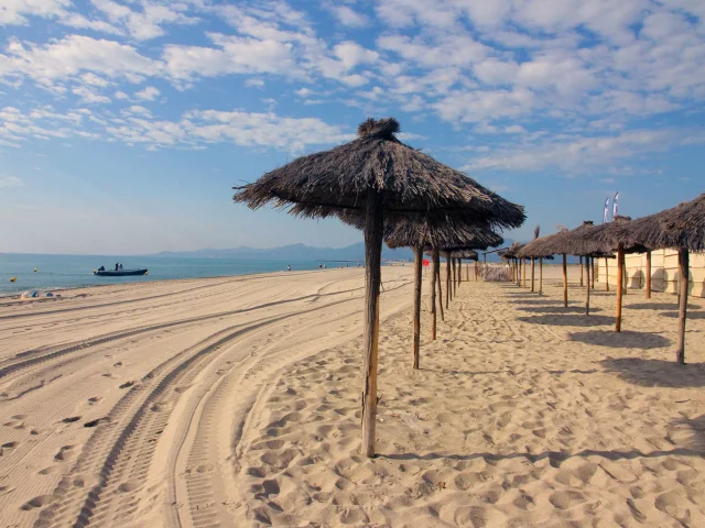 Plage de sable fin bordée de parasols en paille, avec la mer Méditerranée et les montagnes visibles à l’horizon sous un ciel bleu.