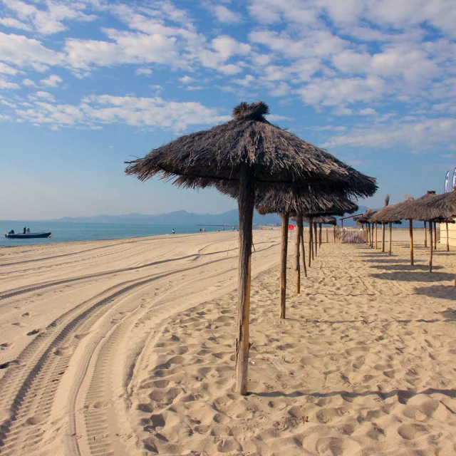 Plage de sable fin bordée de parasols en paille, avec la mer Méditerranée et les montagnes visibles à l’horizon sous un ciel bleu.