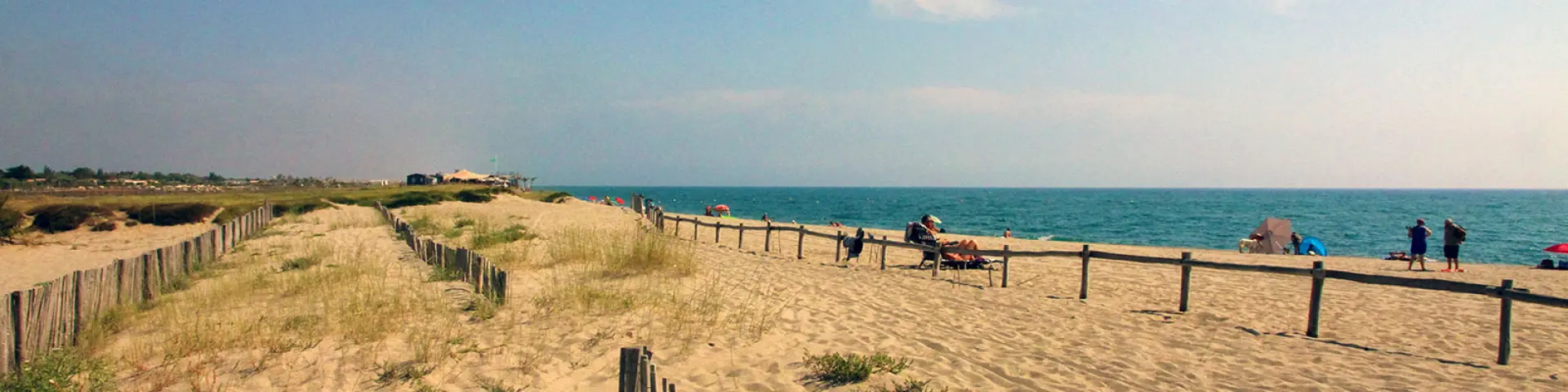 Plage naturelle de Torreilles bordée de dunes et de ganivelles en bois, avec quelques promeneurs et vacanciers face à la mer Méditerranée.