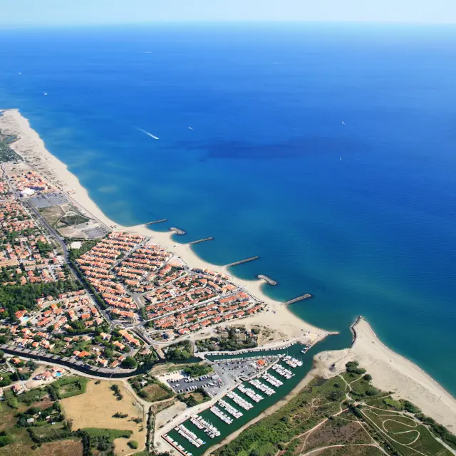 Vue aérienne du littoral des Pyrénées-Orientales avec la station balnéaire, la plage et le port de plaisance bordés par la Méditerranée.