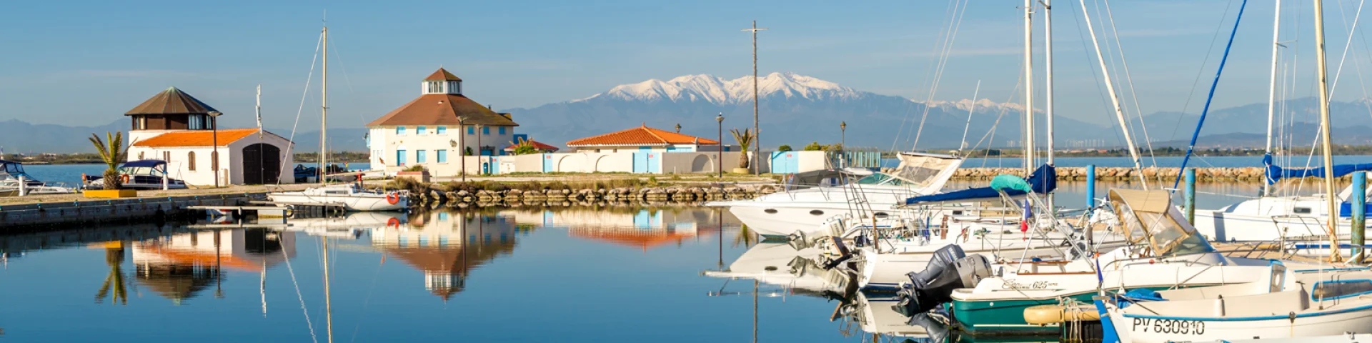Port de plaisance paisible aux eaux calmes, avec voiliers amarrés et le Canigó enneigé en arrière-plan sous un ciel bleu.