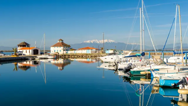 Port de plaisance paisible aux eaux calmes, avec voiliers amarrés et le Canigó enneigé en arrière-plan sous un ciel bleu.