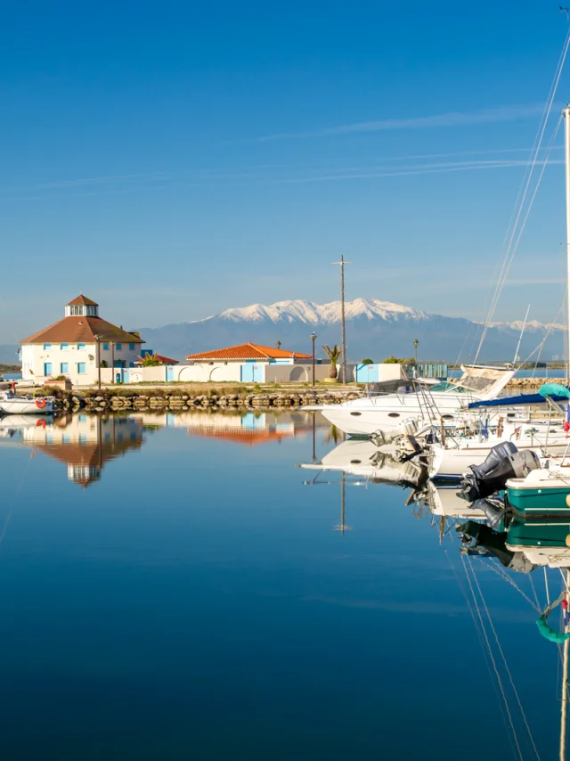 Port de plaisance paisible aux eaux calmes, avec voiliers amarrés et le Canigó enneigé en arrière-plan sous un ciel bleu.