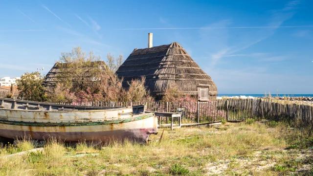 Cabanes traditionnelles en roseaux bordées d’une barque en bois, sur fond de mer bleue et de ciel clair.