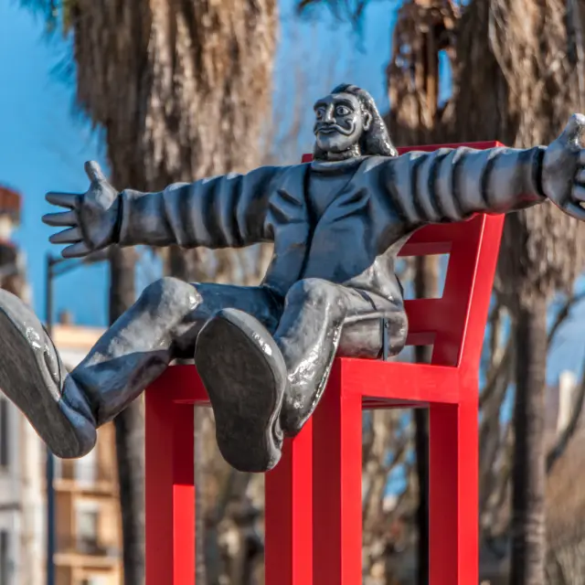 Sculpture contemporaine représentant un homme moustachu assis les bras ouverts sur une grande chaise rouge, en extérieur sous les palmiers.