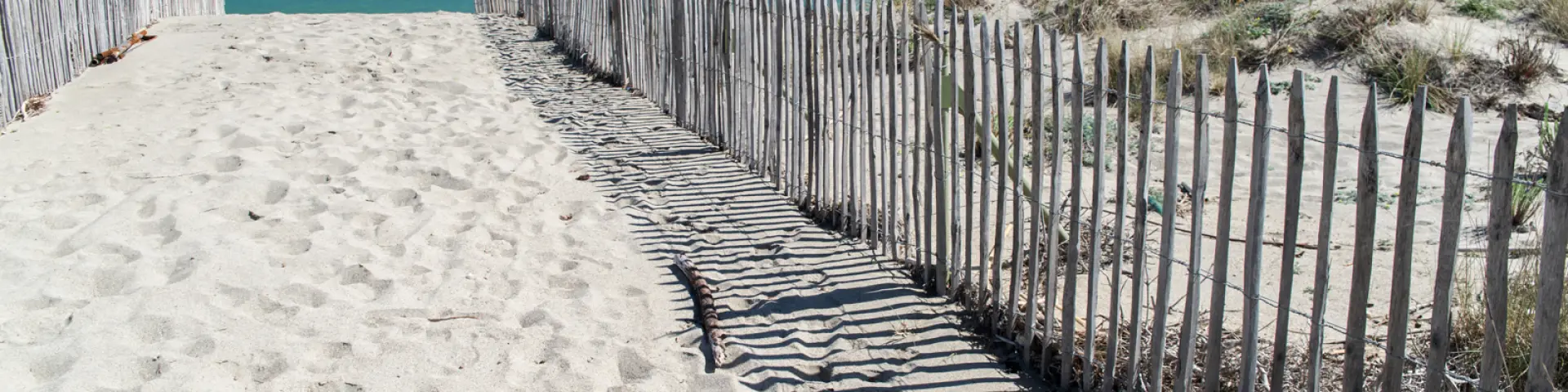 Chemin sablonneux bordé de ganivelles menant à la plage, avec la mer turquoise et le ciel bleu en arrière-plan.