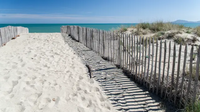 Chemin sablonneux bordé de ganivelles menant à la plage, avec la mer turquoise et le ciel bleu en arrière-plan.
