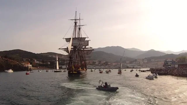 Arrivée de l'Hermione à Port-Vendres. Grand voilier à trois mâts entrant dans le port de Port-Vendres, entouré de petites embarcations et de spectateurs massés sur les quais au coucher du soleil.