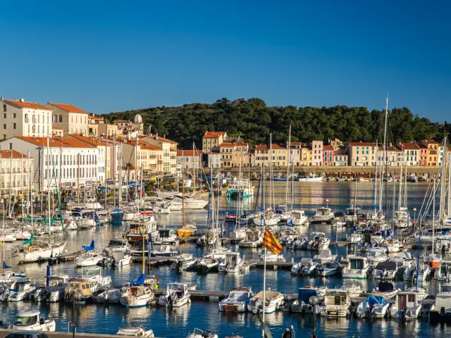 Vue panoramique du port de plaisance animé, bordé de façades colorées aux toits de tuiles rouges, sous un ciel bleu limpide.