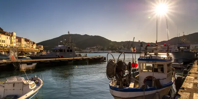 Bateaux de pêche amarrés dans le port au coucher du soleil, avec les collines en arrière-plan et les façades colorées du front de mer.