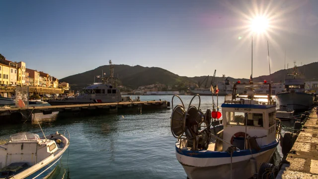 Bateaux de pêche amarrés dans le port au coucher du soleil, avec les collines en arrière-plan et les façades colorées du front de mer.