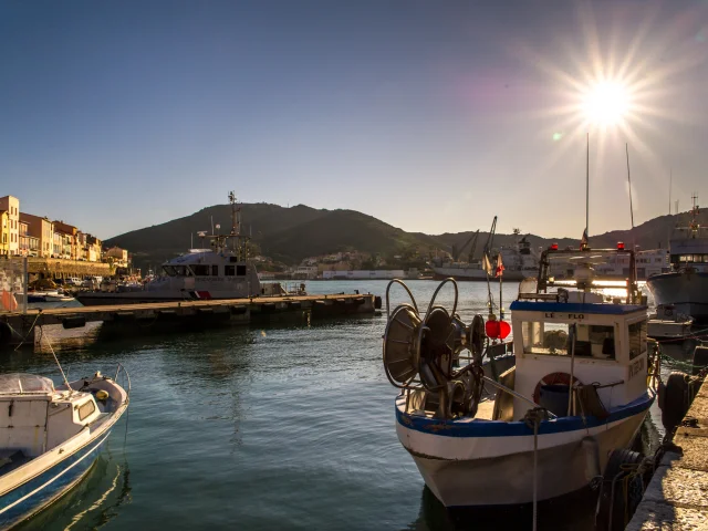 Bateaux de pêche amarrés dans le port au coucher du soleil, avec les collines en arrière-plan et les façades colorées du front de mer.