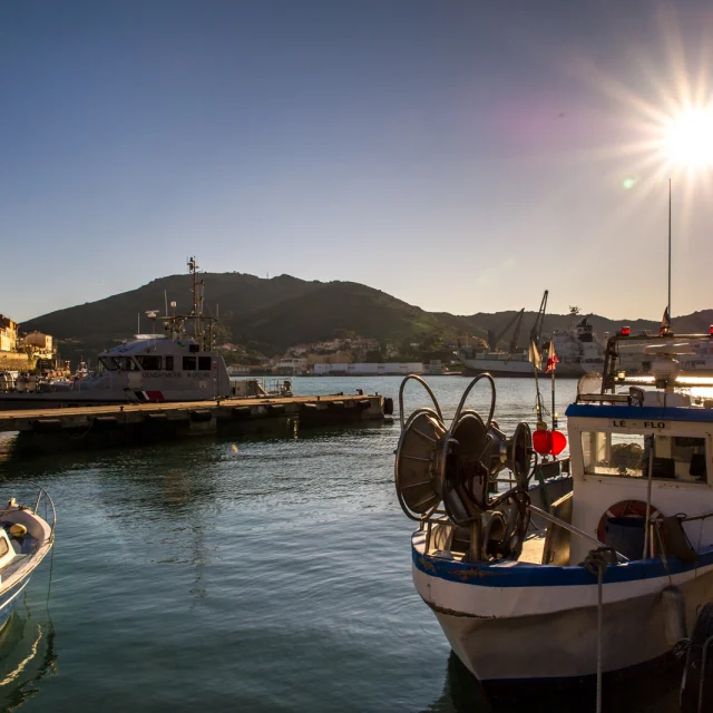 Bateaux de pêche amarrés dans le port au coucher du soleil, avec les collines en arrière-plan et les façades colorées du front de mer.