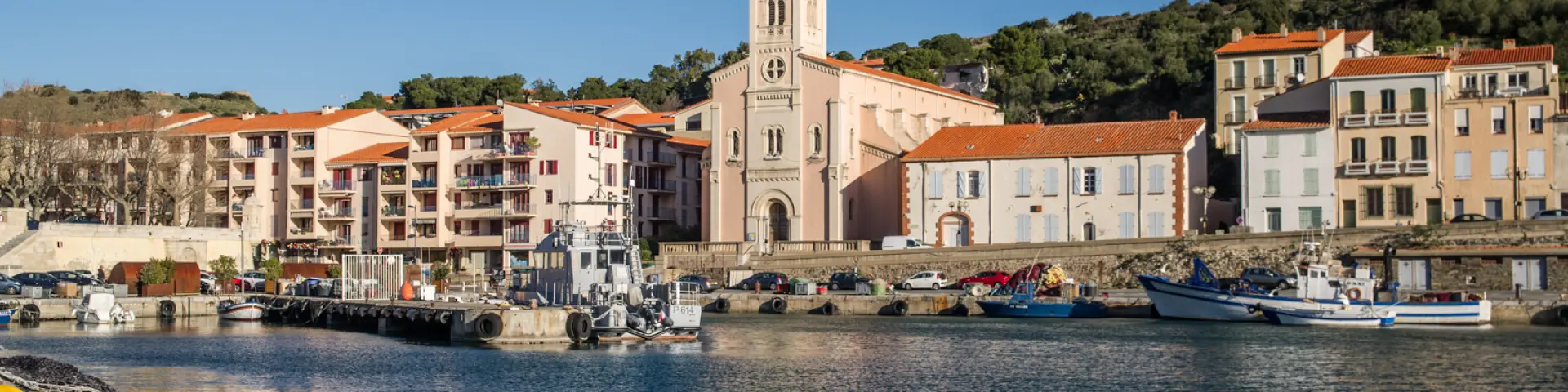Vue du port de plaisance et de pêche bordé d’immeubles pastel et d’une église à coupole blanche sous un ciel bleu éclatant.