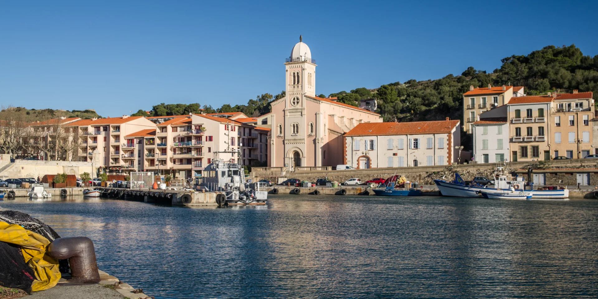 Vue du port de plaisance et de pêche bordé d’immeubles pastel et d’une église à coupole blanche sous un ciel bleu éclatant.