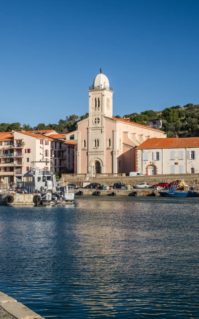 Vue du port de plaisance et de pêche bordé d’immeubles pastel et d’une église à coupole blanche sous un ciel bleu éclatant.