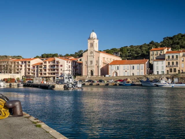 Vue du port de plaisance et de pêche bordé d’immeubles pastel et d’une église à coupole blanche sous un ciel bleu éclatant.