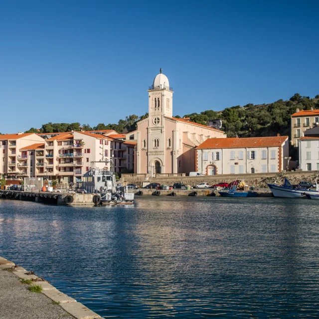 Vue du port de plaisance et de pêche bordé d’immeubles pastel et d’une église à coupole blanche sous un ciel bleu éclatant.