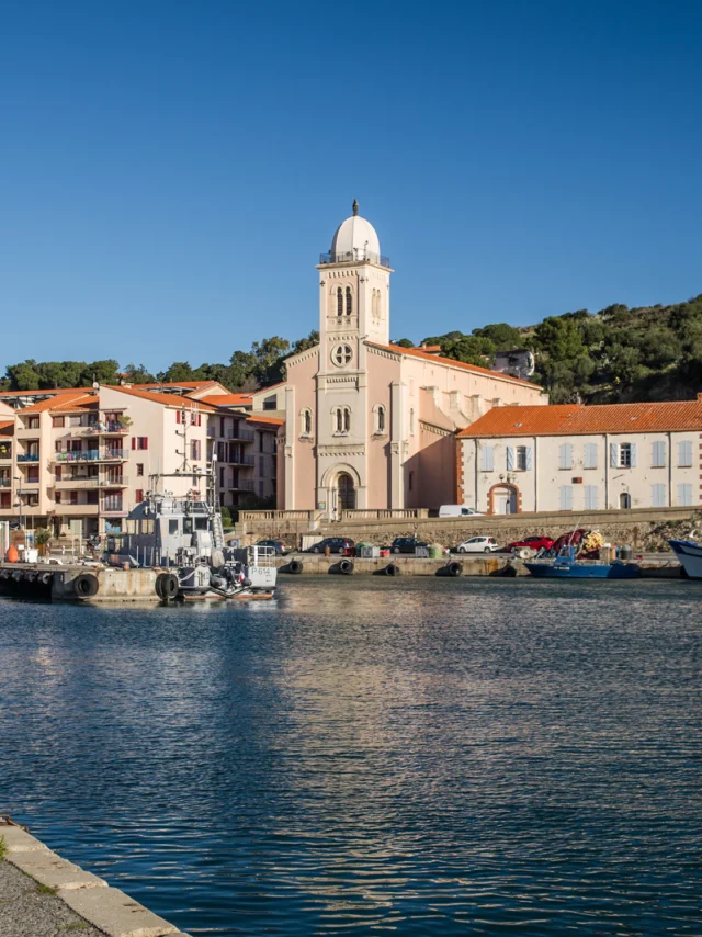 Vue du port de plaisance et de pêche bordé d’immeubles pastel et d’une église à coupole blanche sous un ciel bleu éclatant.