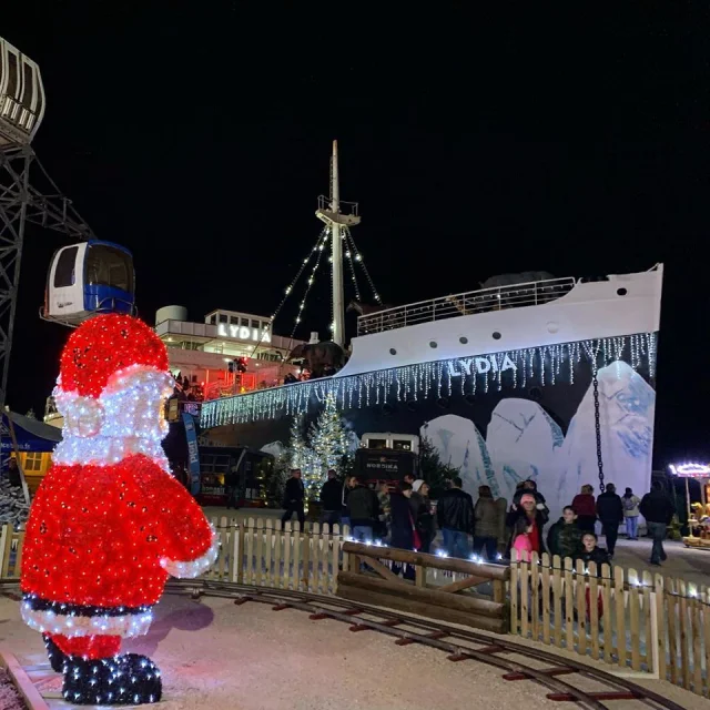 Décor de Noël au Barcarès avec le paquebot Lydia illuminé, des guirlandes scintillantes, un grand Père Noël lumineux et des visiteurs se promenant de nuit.