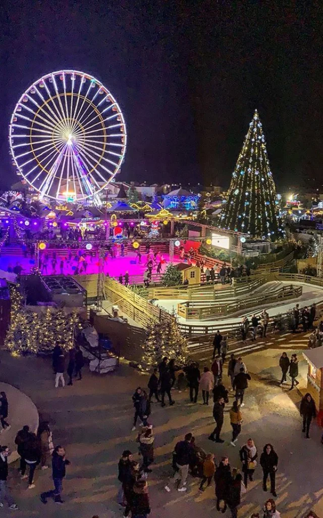 Village de Noël du Barcarès de nuit avec grande roue illuminée, patinoire, sapin géant décoré et foule de visiteurs profitant des animations.