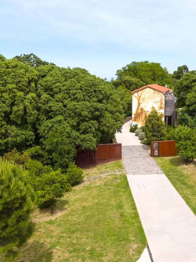 Vue aérienne de l’entrée du site des anciennes dynamiteries Alfred Nobel à Paulilles. Un chemin pavé traverse un espace verdoyant menant à un bâtiment restauré, orné d’une fresque murale représentant des ouvriers du site, entouré d’arbres et de végétation dense.