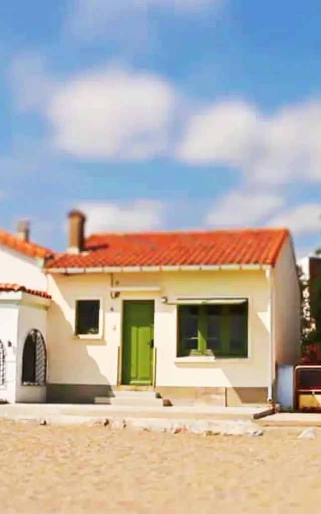 Alignement de petites maisons colorées du Racou à Argelès-sur-Mer, photographiées de face depuis la plage, avec leurs toits en tuiles rouges, façades pastel et volets colorés sous un ciel bleu légèrement nuageux.