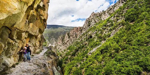 Deux randonneuses avancent prudemment sur un sentier étroit taillé dans la falaise, longeant une paroi rocheuse vertigineuse des Gorges de la Carança, entourée de montagnes verdoyantes sous un ciel partiellement nuageux.
