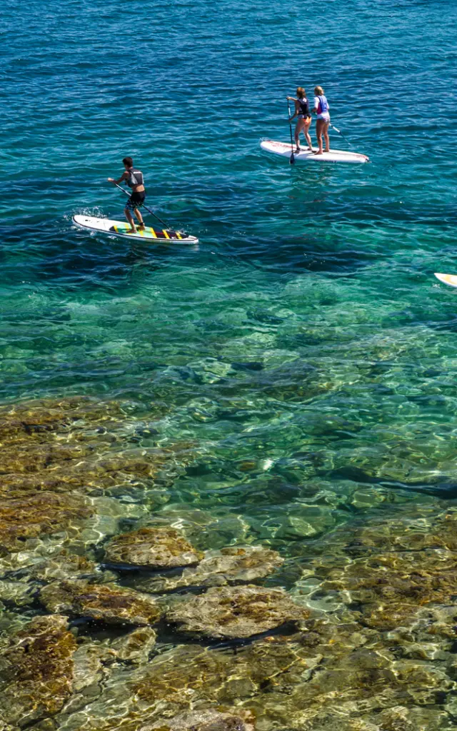 Groupe de personnes pratiquant le paddle sur une mer turquoise et transparente, au-dessus d’un fond rocheux visible.