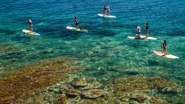 Groupe de personnes pratiquant le paddle sur une mer turquoise et transparente, au-dessus d’un fond rocheux visible.