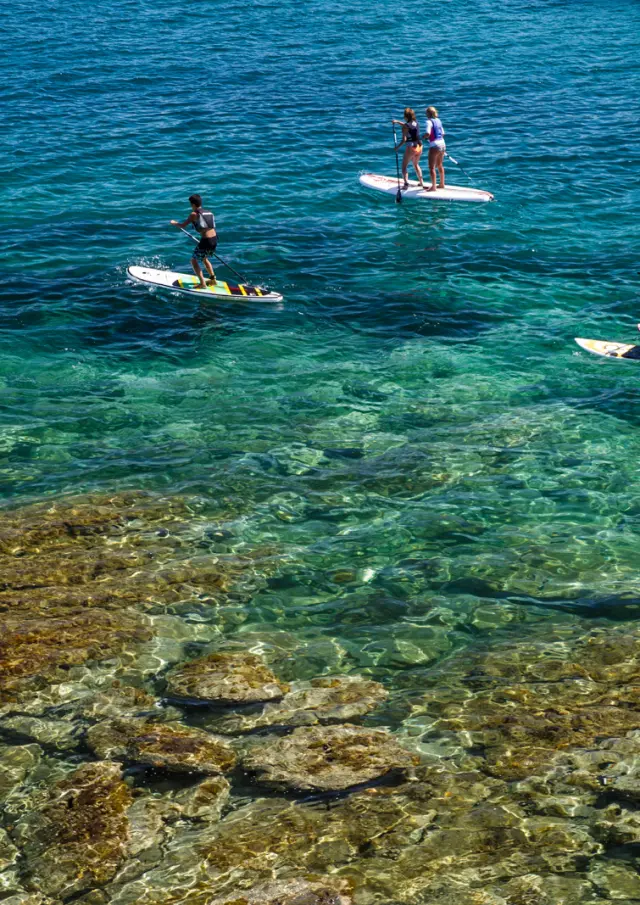 Groupe de personnes pratiquant le paddle sur une mer turquoise et transparente, au-dessus d’un fond rocheux visible.