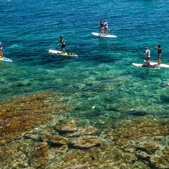 Groupe de personnes pratiquant le paddle sur une mer turquoise et transparente, au-dessus d’un fond rocheux visible.